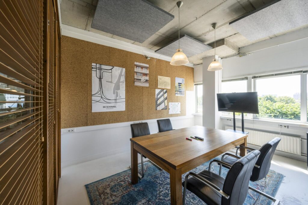 Vergaderruimte met houten tafel, vier zwarte stoelen, een prikbord met architectuurposters en een scherm bij het raam aan de Anthony Fokkerweg.